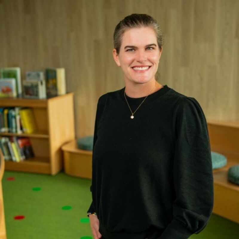 The image shows a woman with short hair, wearing a black sweater and a necklace, smiling at the camera. She is standing in a library with bookshelves and a green carpet. The background suggests a well-lit and inviting space, possibly a school or community library.